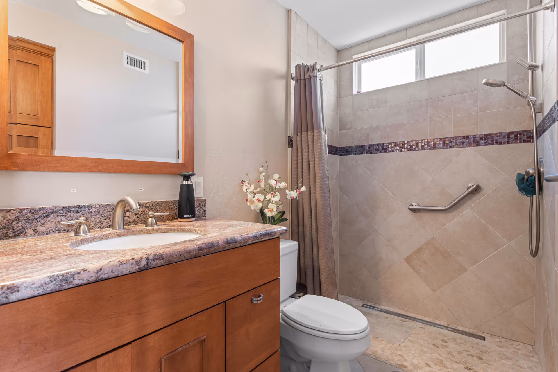 A bathroom with a wooden vanity featuring a granite countertop and an undermount sink. Above the vanity is a large wooden-framed mirror. To the right is a toilet with a vase of white and pink flowers placed on top of the tank. Next to the toilet is a walk-in shower with beige tiled walls, a horizontal strip of dark mosaic tiles, a grab bar, and a handheld showerhead. A window above the shower allows natural light into the space.