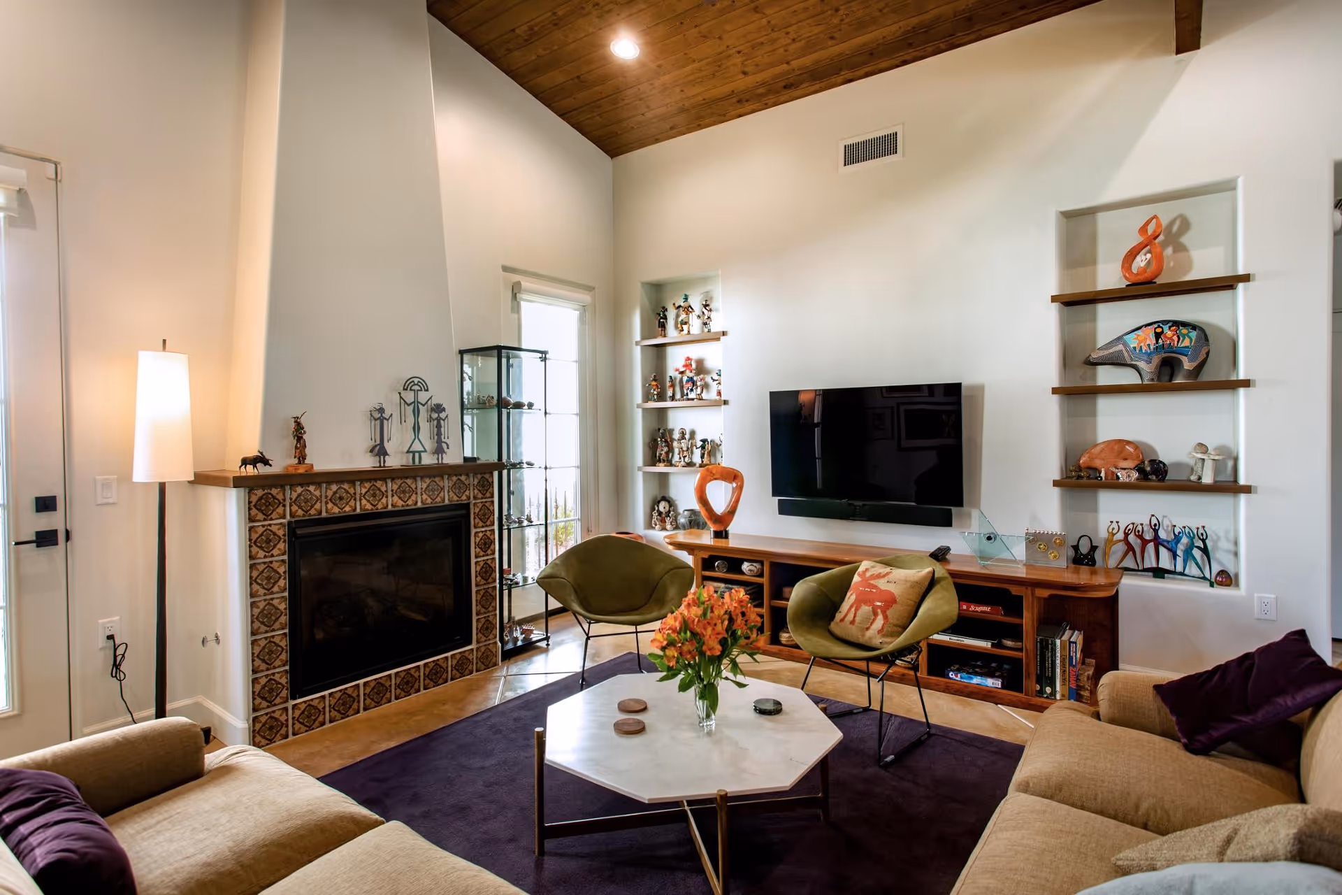 A cozy living room with a high wooden ceiling, a tiled fireplace, two green chairs, a beige sofa with purple pillows, a hexagonal white coffee table with a vase of orange flowers, a wall-mounted flat screen TV, and shelves displaying various decorative items and figurines.