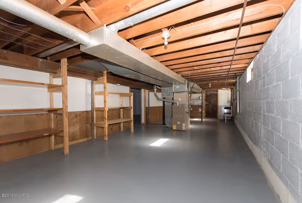 Unfinished basement with a painted concrete floor, exposed ceiling joists, ductwork, shelving and utility equipment.