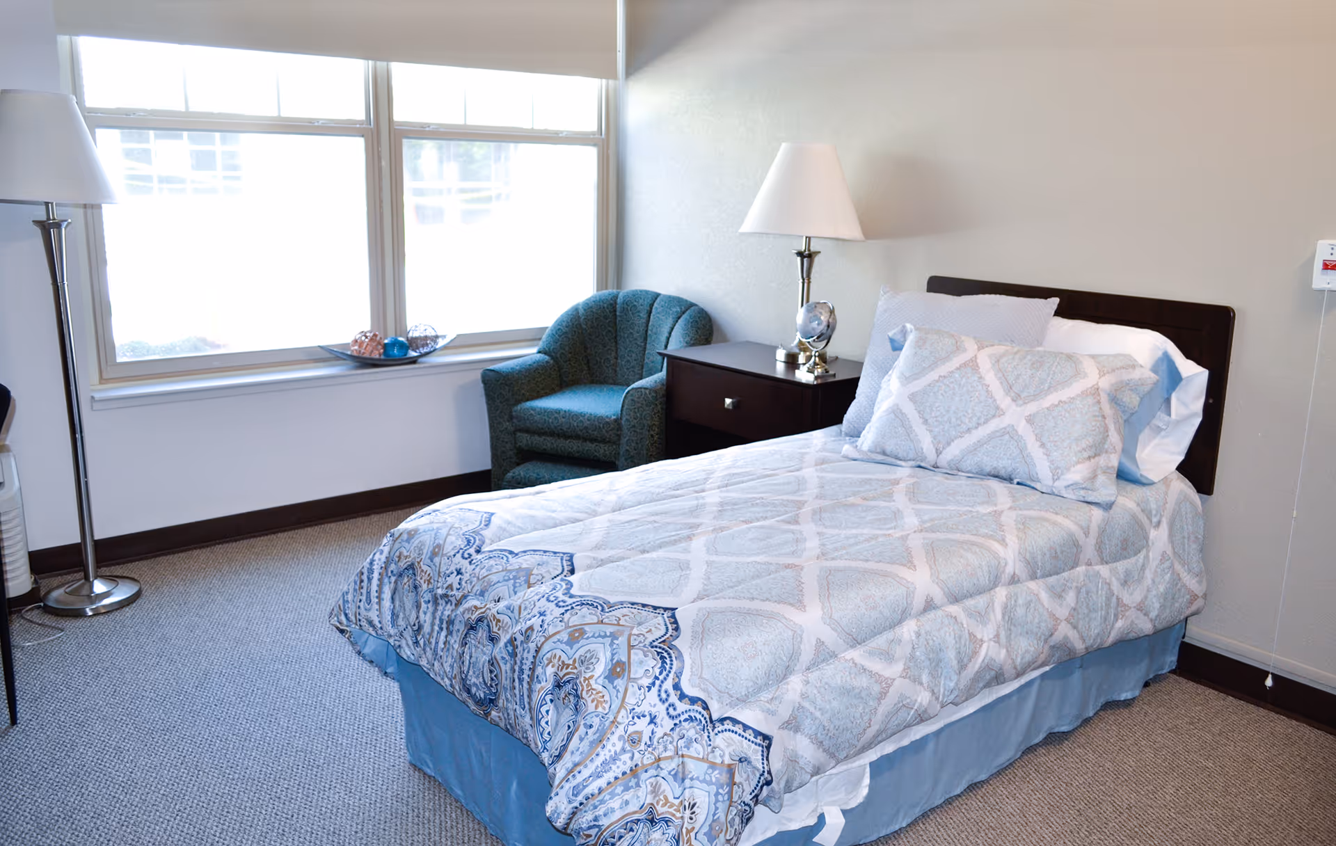 A bright single bedroom with a made bed, nightstand, armchair and large window.