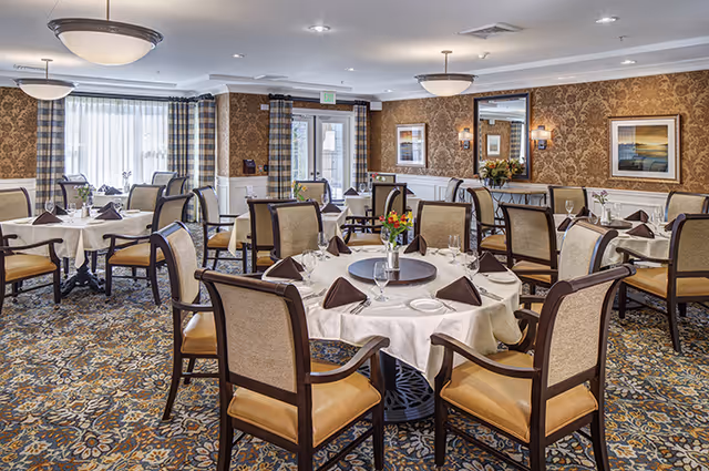 A well-lit dining room with multiple round tables covered in white tablecloths, each set with plates, glasses, napkins folded in a triangle, and small flower arrangements. The room features patterned carpet, wallpaper with a floral design, framed artwork on the walls, and large windows with curtains allowing natural light to enter.