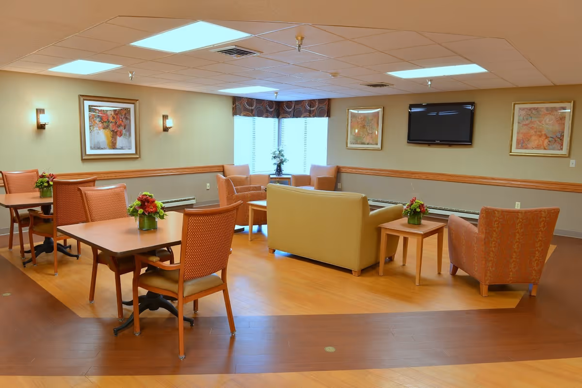 A bright senior living common room with dining tables and chairs, lounge seating, wall art and a wall-mounted TV.