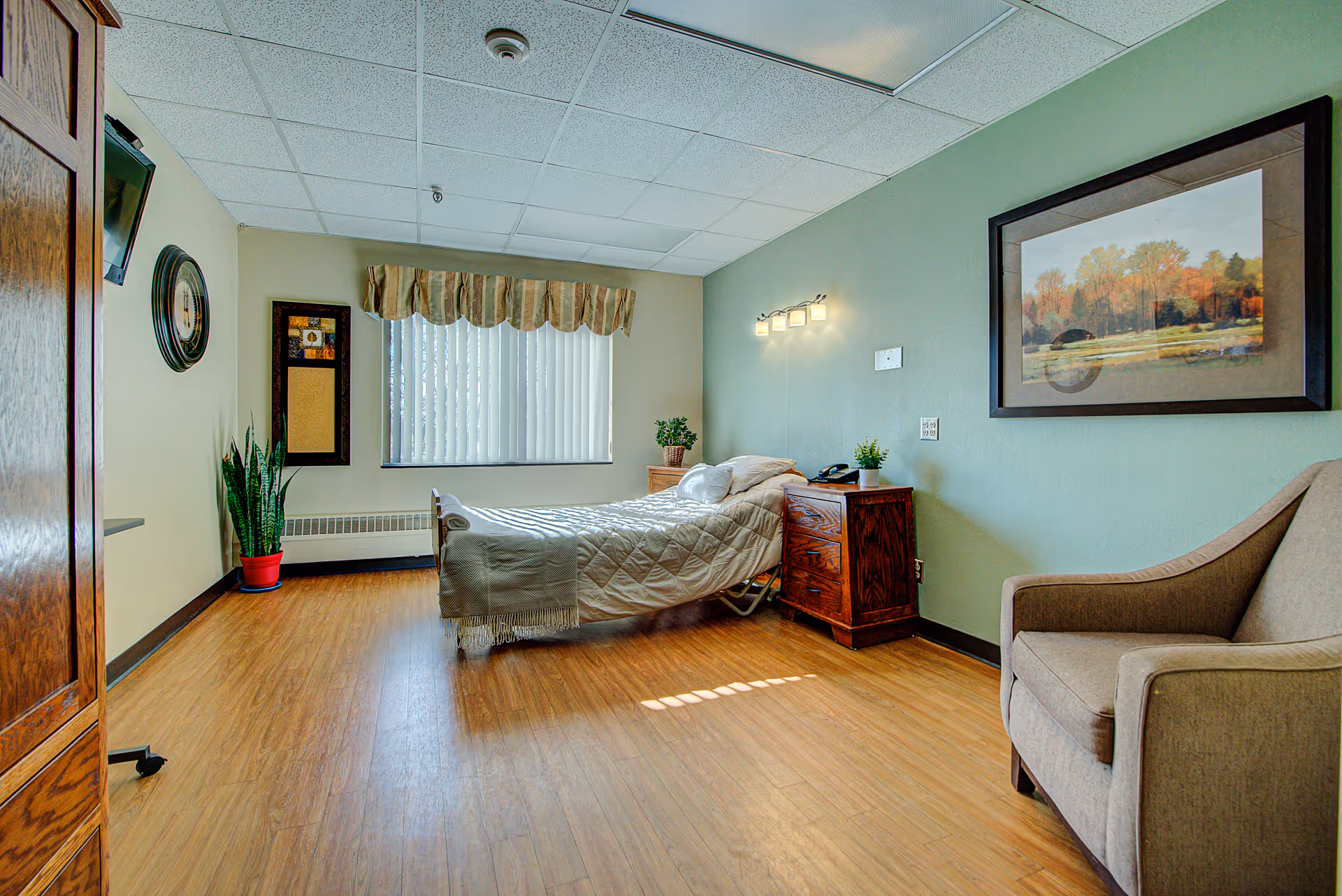 A bright and clean bedroom in a senior living facility with a single bed covered in a beige quilt, a wooden nightstand with a phone and plant, a comfortable armchair, a large window with vertical blinds and a valance, a wall-mounted TV, a clock, and framed artwork on the walls. The floor is wood laminate and the walls are painted light green and beige.