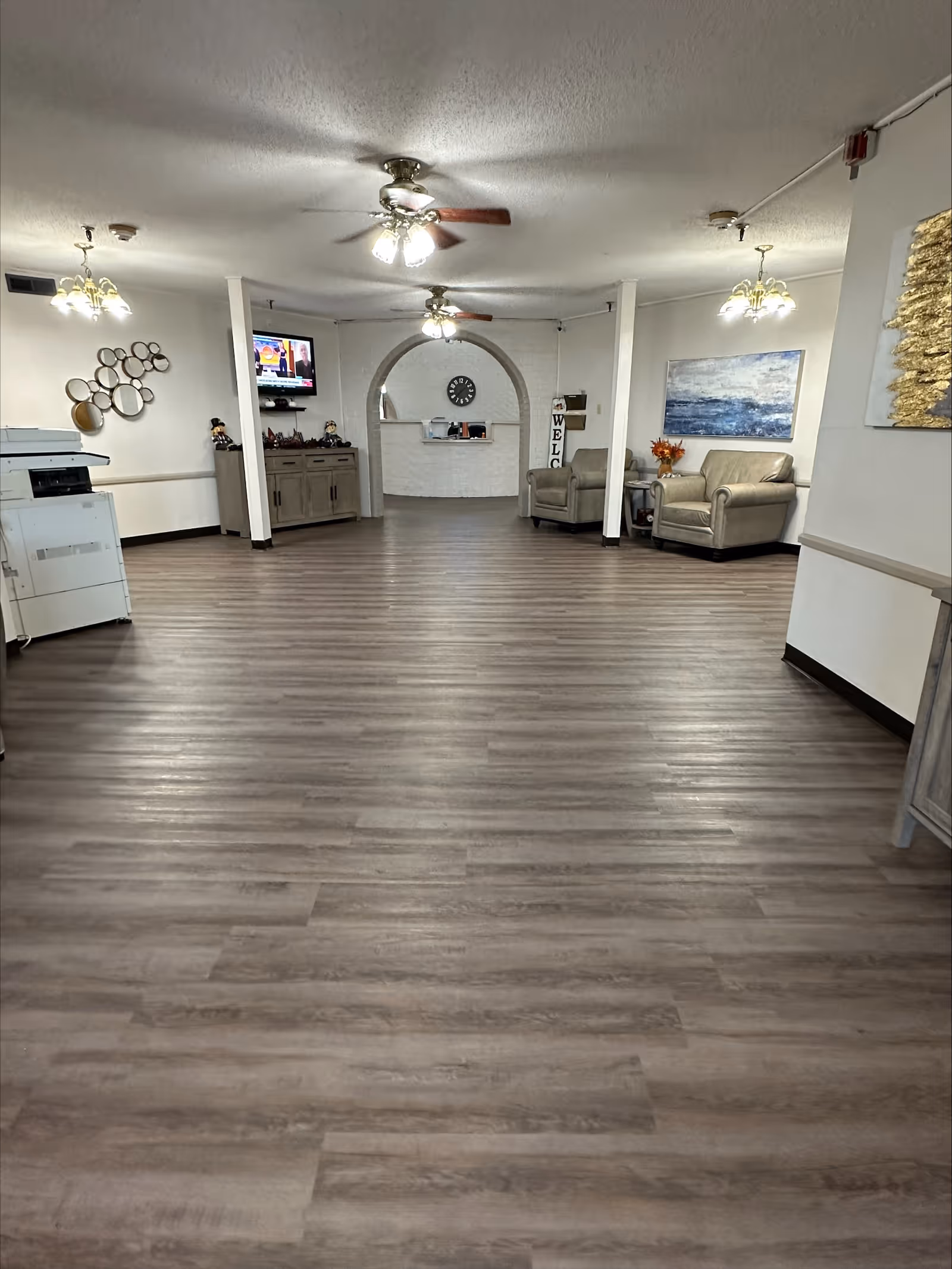 Interior view of a nursing and rehab facility common area with wood flooring, two ceiling fans with lights, two beige armchairs, a wall-mounted TV, decorative wall art, and a clock above an arched opening in a white brick wall.