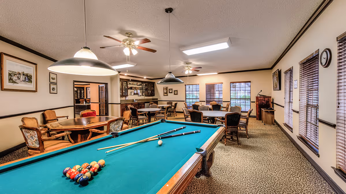 A spacious recreation room with a pool table in the foreground, complete with billiard balls and cues. The room features multiple round tables with chairs, ceiling fans, pendant lights, and large windows with blinds letting in natural light. The walls are decorated with framed pictures and a clock.