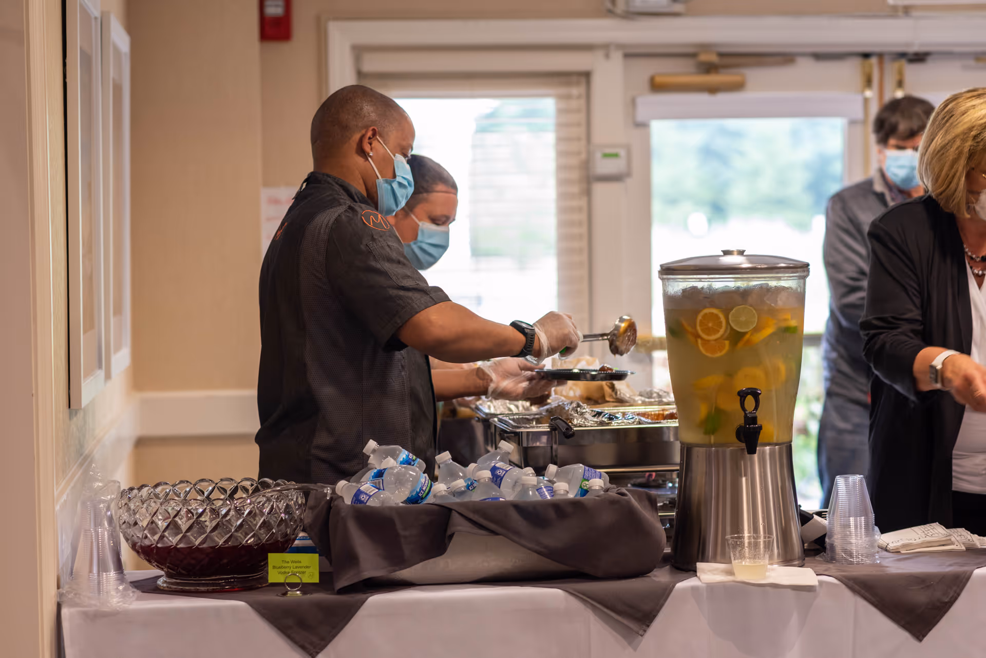Two masked staff members serving food from chafing dishes at a buffet table with bottled water and a large beverage dispenser filled with citrus-infused water, while other masked people stand nearby in a well-lit room.