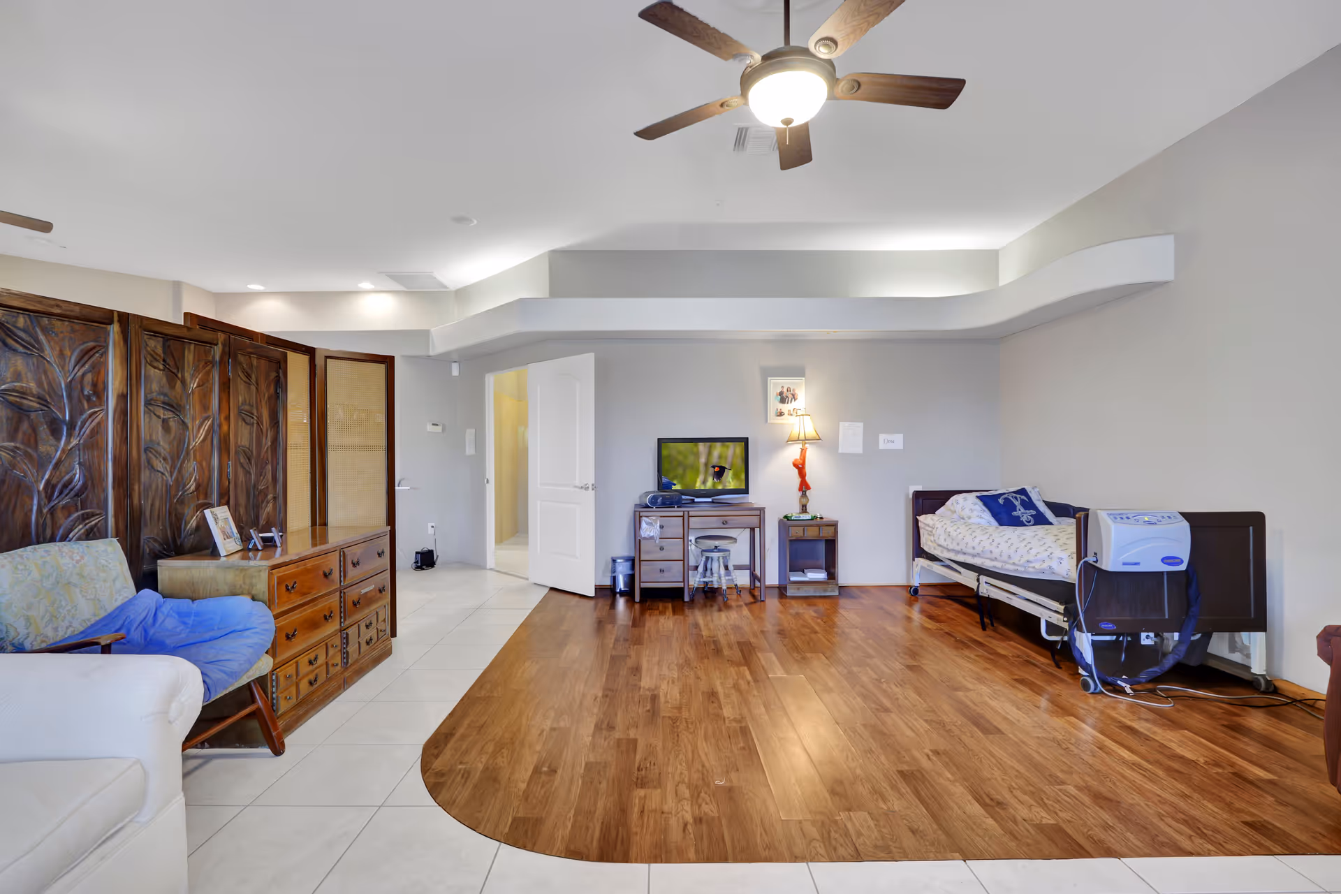 A senior living facility room with a single bed equipped with medical devices on the right side, a small wooden desk with a TV and a lamp on it, a wooden dresser, a cushioned chair with a blue blanket, and a decorative wooden room divider. The floor is a combination of tile and wood, and there is a ceiling fan with a light fixture.