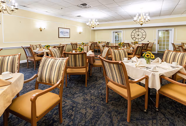 A dining room with multiple tables covered with beige tablecloths, each set with white napkins, cups, and small vases holding sunflowers. The room has striped cushioned chairs, chandeliers hanging from the ceiling, a large decorative clock on the far wall, and framed artwork on the side wall.