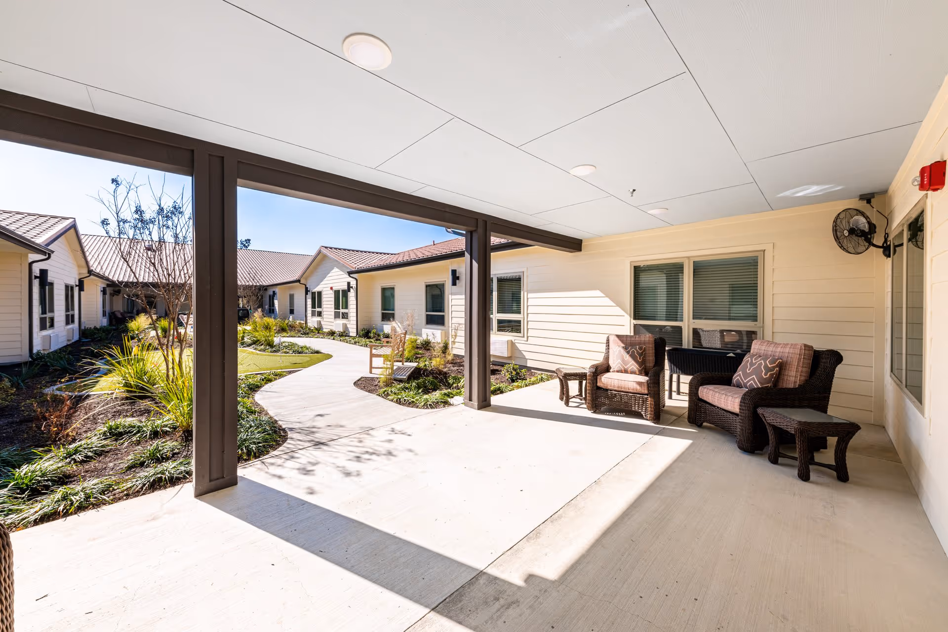 Covered outdoor patio area with wicker chairs and small tables, adjacent to a walkway surrounded by landscaped garden beds and single-story building units with windows and beige siding under a clear blue sky.