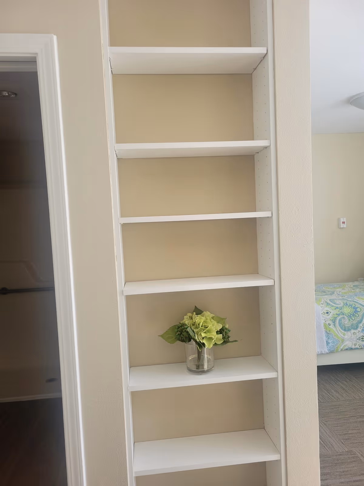 A white built-in shelving unit with six shelves, one of which holds a small glass vase with green flowers. To the left, there is a doorway leading to a bathroom with a grab bar visible. To the right, part of a bedroom with a bed covered in a green and blue patterned bedspread is visible.