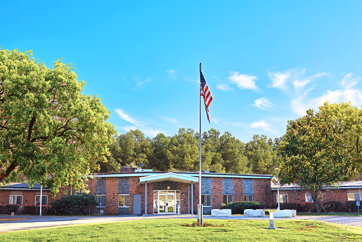 Front exterior view of The Grand Rehabilitation and Nursing at Guilderland building with a flagpole displaying the American flag in the foreground, surrounded by green trees and a clear blue sky.
