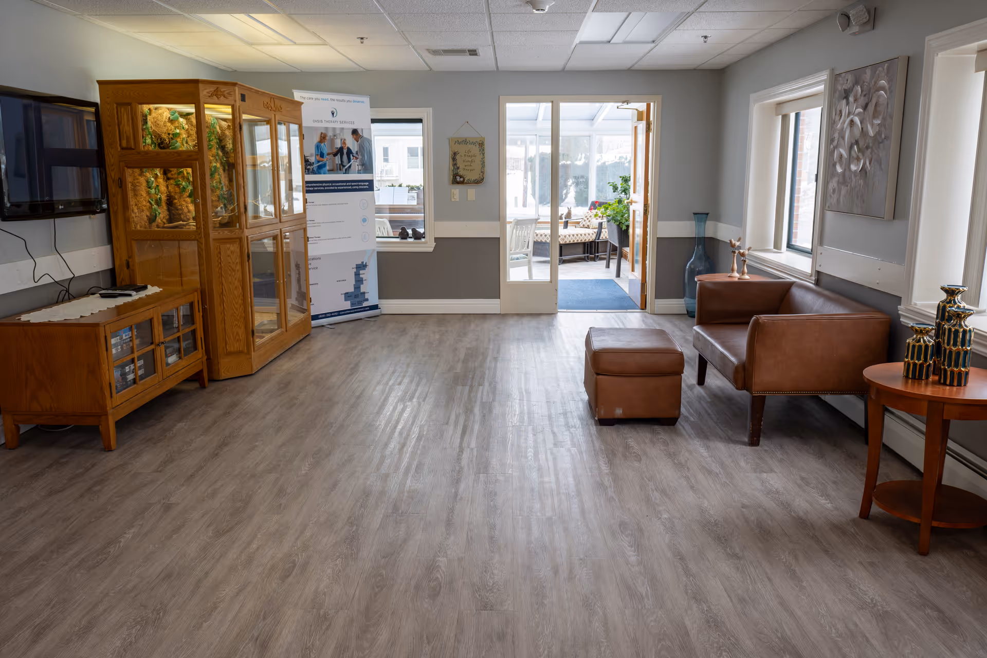 A spacious, well-lit common area with wood flooring, a brown leather loveseat with matching ottoman, a wooden side table with decorative vases, a wooden cabinet with glass doors, a wall-mounted TV, and a glass door leading to a sunroom with seating and plants.