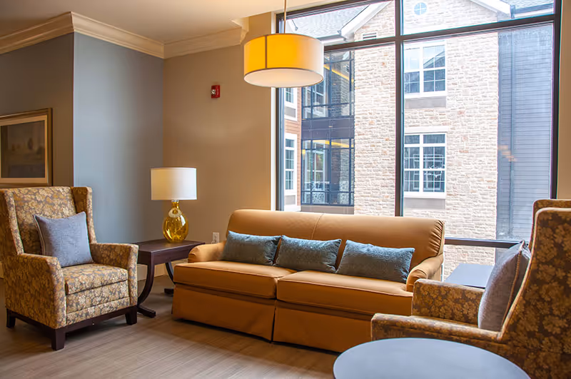 Cozy seating area with a sofa, armchairs, side tables, and a lamp in front of large windows overlooking an adjacent building.