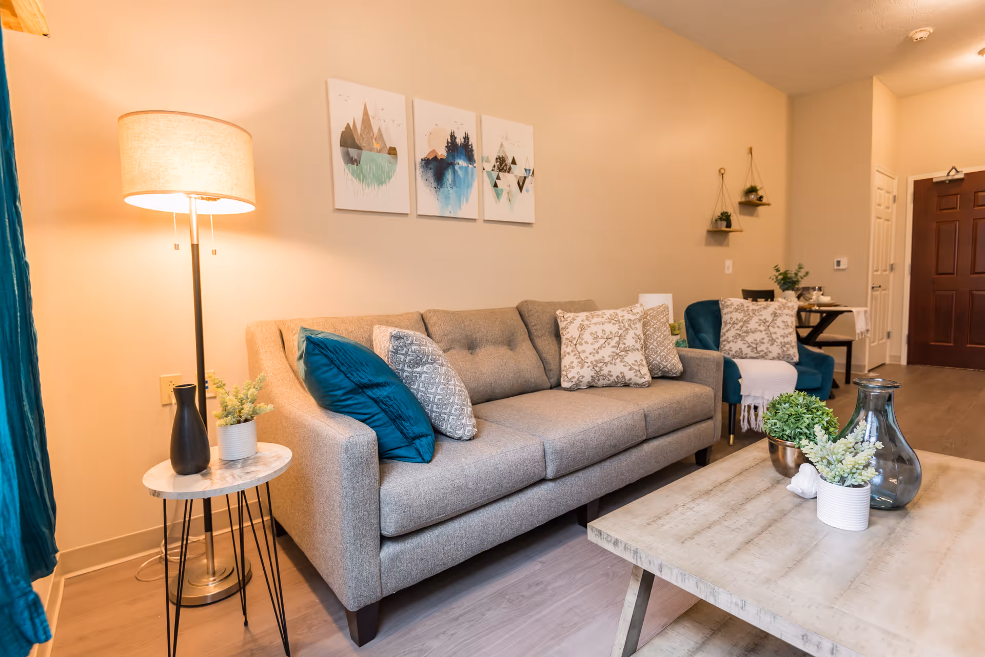 A cozy living room with a gray sofa adorned with patterned and solid teal pillows, a small round side table with a lamp and decorative vases, a wooden coffee table with potted plants and a glass vase, teal armchairs with pillows, wall art featuring abstract mountain and water designs, and a dark wooden door in the background.