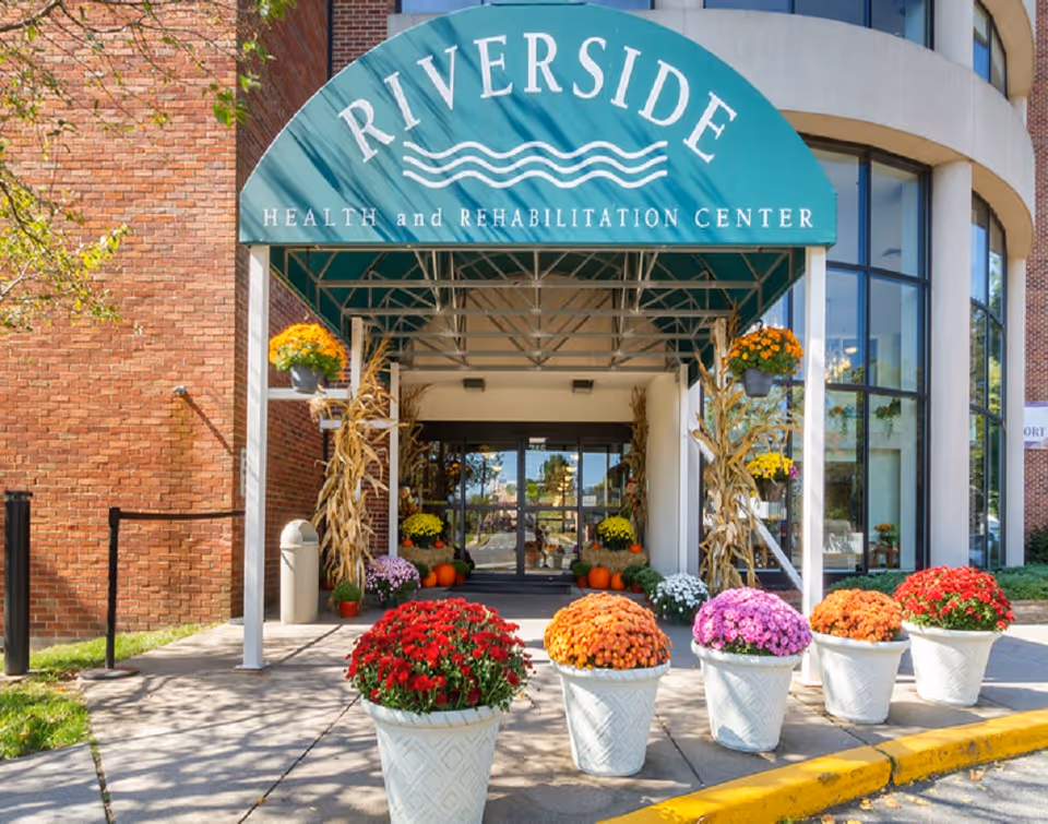 Entrance to Riverside Health and Rehabilitation Center with a green awning displaying the facility name. The entrance is decorated with colorful potted flowers and autumn-themed decorations including cornstalks and pumpkins. The building has large windows and a brick exterior.