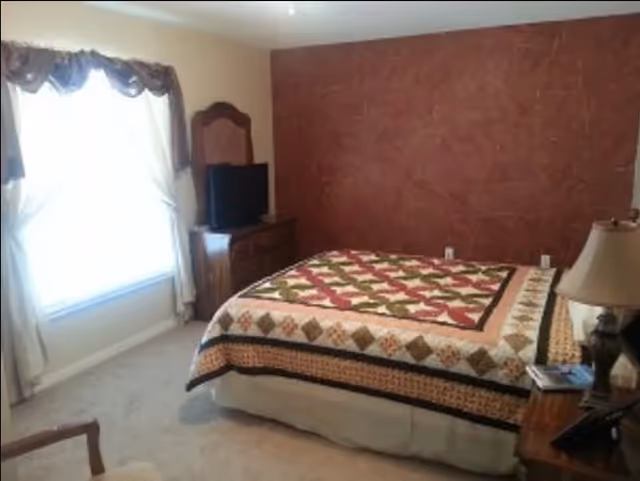 A bedroom featuring a bed with a patchwork quilt, a dresser-mounted TV, a nightstand with a lamp, and a curtained window.
