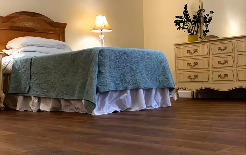 Neatly made bed with a green quilt, bedside lamp, and a dresser in a warmly lit bedroom with wood flooring.