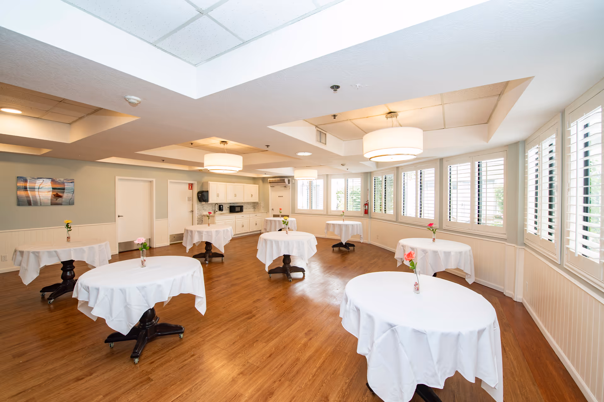 Bright dining room with several small round tables covered in white tablecloths, hardwood floors, and large windows with shutters.