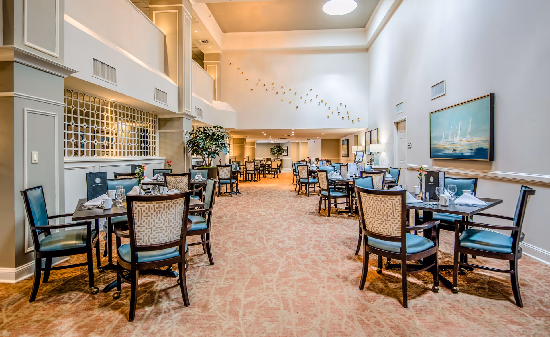 A spacious and well-lit dining room with multiple tables and chairs arranged neatly. The room features high ceilings, a skylight, decorative wall art, and a carpeted floor. Tables are set with glassware, napkins, and menus, creating an inviting atmosphere.