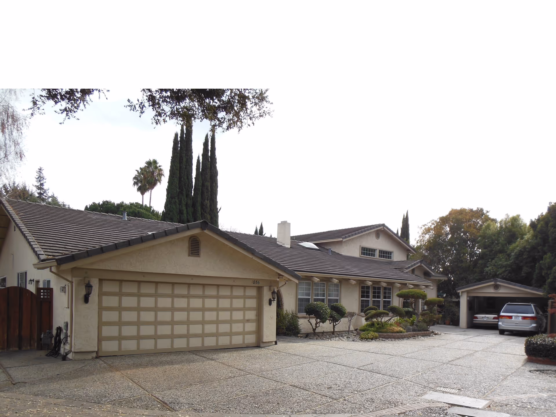 Exterior view of a residential building with a large driveway, two garages, and well-maintained landscaping including trimmed bushes and tall trees in the background.