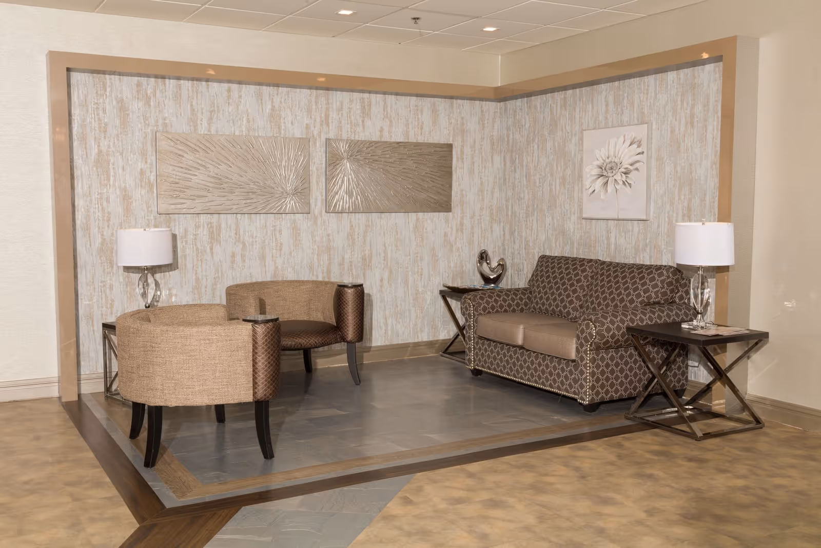 A cozy seating area in a healthcare facility featuring a patterned brown loveseat, two beige armchairs, two side tables with glass lamps, and decorative wall art including metallic abstract pieces and a floral painting.