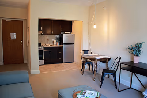 A small living space featuring a kitchenette with dark cabinets, a refrigerator, and a stove. Adjacent to the kitchenette is a wooden dining table with two black metal chairs. A light bulb hangs from the ceiling above the table. There is a blue sofa and ottoman in the foreground, and a black console table with a potted plant on the right side. The entrance door is visible in the background.