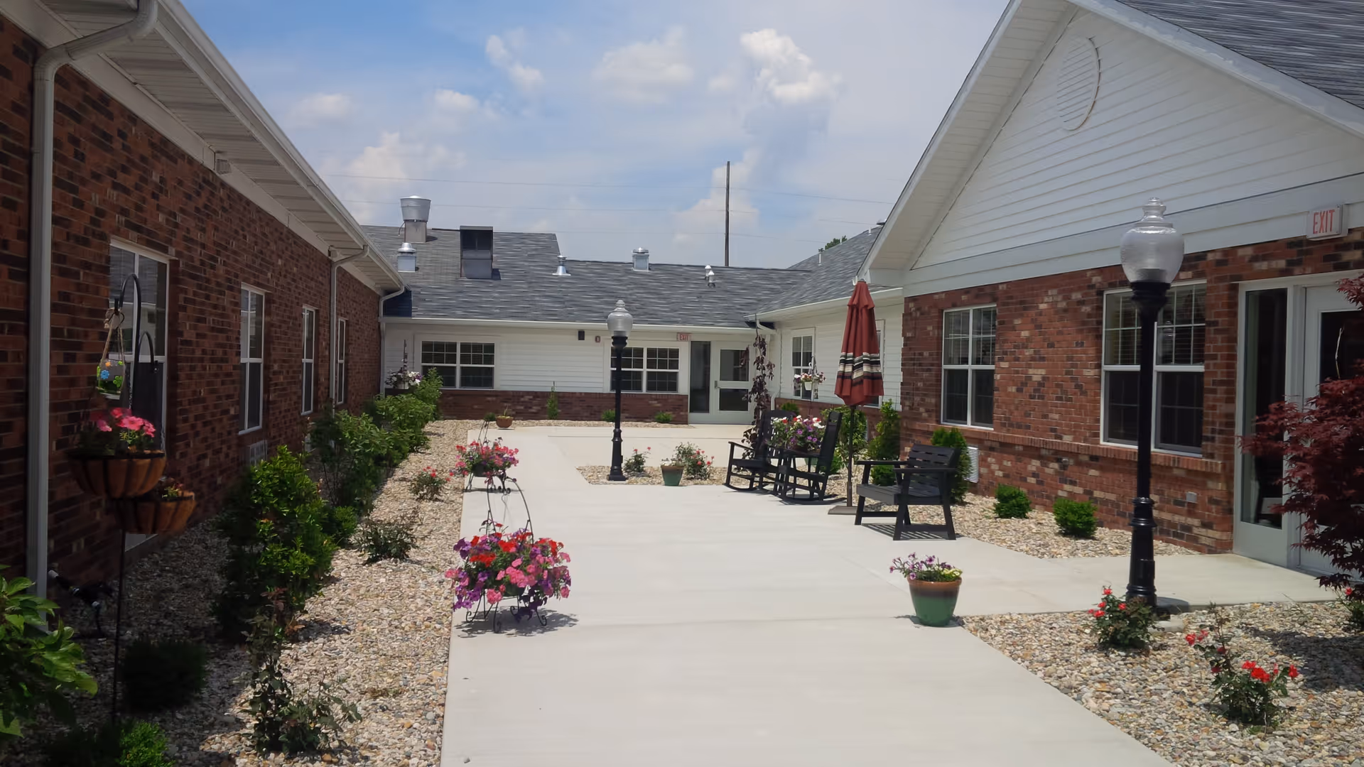 Outdoor courtyard area of Amber Manor Care Center with brick and white siding buildings on either side, a concrete walkway, flower pots with colorful flowers, benches, and lamp posts under a partly cloudy sky.