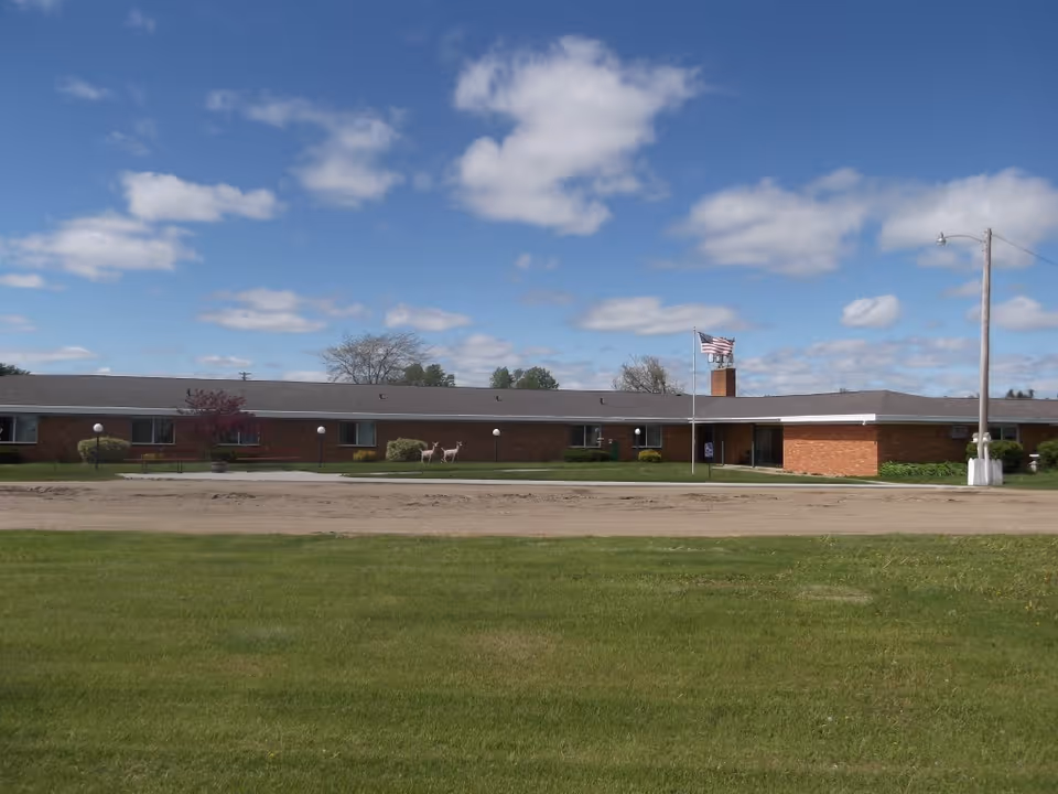 Single-story brick building with a flat roof under a partly cloudy blue sky. There is a green lawn in the foreground, a dirt road or path in front of the building, and an American flag flying on a flagpole near the entrance. Some small bushes and a tree are planted near the building.