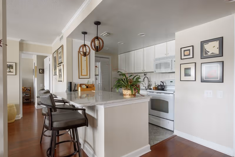Interior view of a kitchen area with white cabinets, a white stove, and a microwave. A marble countertop island with two cushioned bar stools is in the foreground. Two pendant lights hang above the island. There is a plant in a basket on the countertop. The walls are decorated with framed artwork, and the floor is wooden.