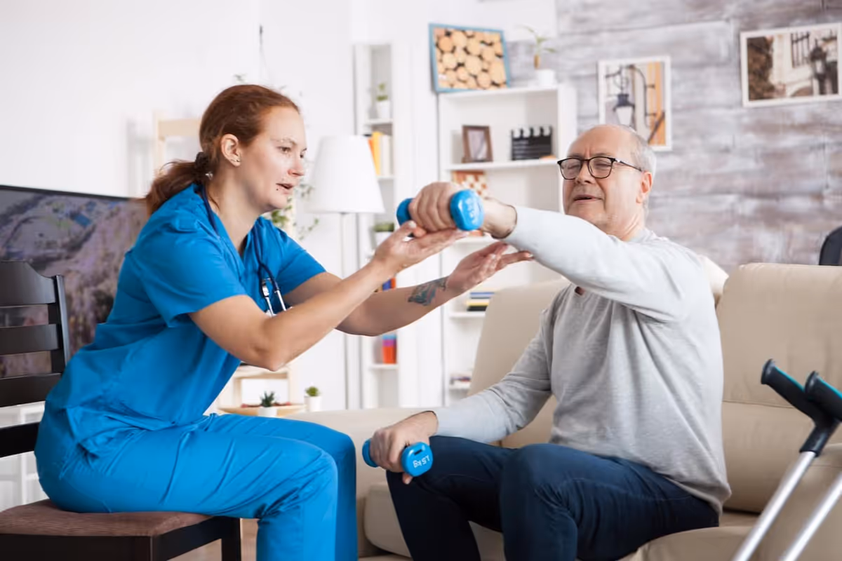A healthcare professional in blue scrubs assists an elderly man sitting on a beige couch with arm exercises using blue dumbbells in a well-lit living room setting.