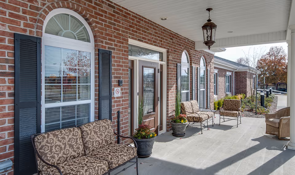 Covered outdoor patio area of a senior living facility with brick walls, arched windows with black shutters, cushioned metal chairs and a wicker chair, potted plants, and a hanging lantern light fixture.