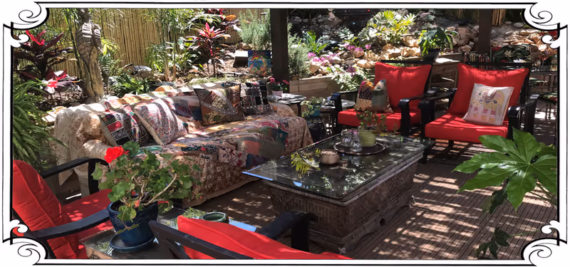 Outdoor patio area with cushioned seating including a sofa covered with a patterned quilt and several red cushioned chairs surrounding a glass-top coffee table. The space is decorated with potted plants and surrounded by lush greenery and flowers.