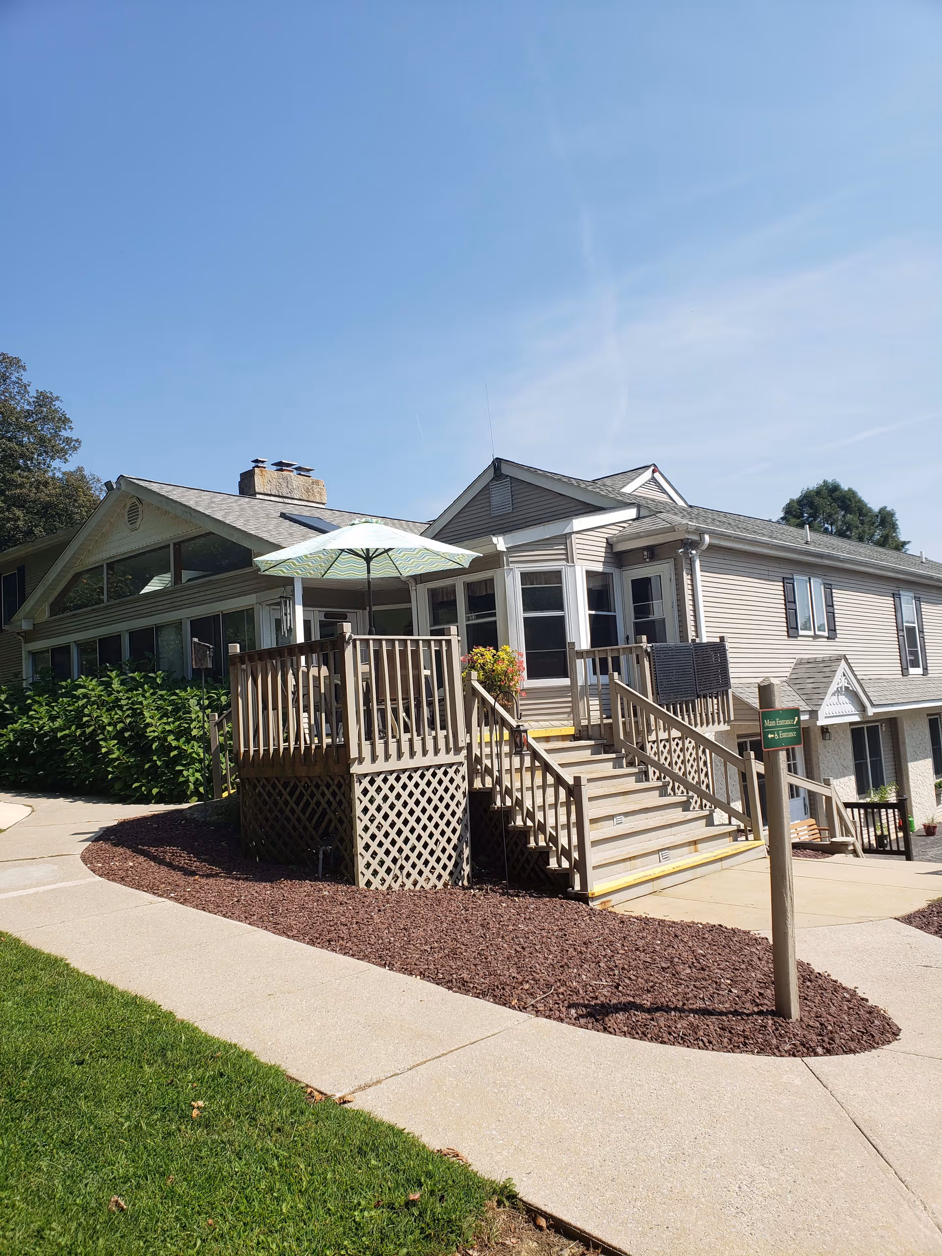 Exterior view of a light-colored building with a wooden deck and stairs leading up to the entrance. The deck has a patio umbrella and some outdoor furniture. There is a paved walkway and landscaped area with mulch and green grass surrounding the building. The sky is clear and blue.