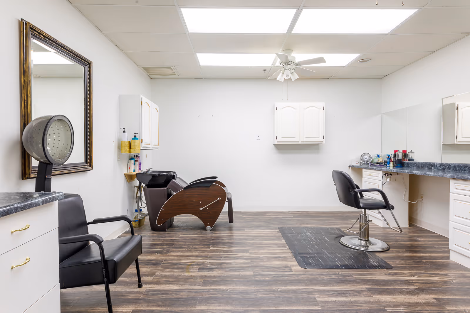 Interior of a senior living facility hair salon with a black salon chair in front of a large mirror, a hair washing station with a reclining chair, white cabinets, and wooden flooring.
