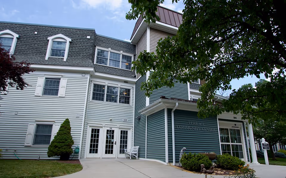 Exterior view of a senior living facility building named Terrace Community Center at Westminster Woods. The building has white and green siding with multiple windows, a white door entrance, and a small garden area with shrubs and a tree in front.