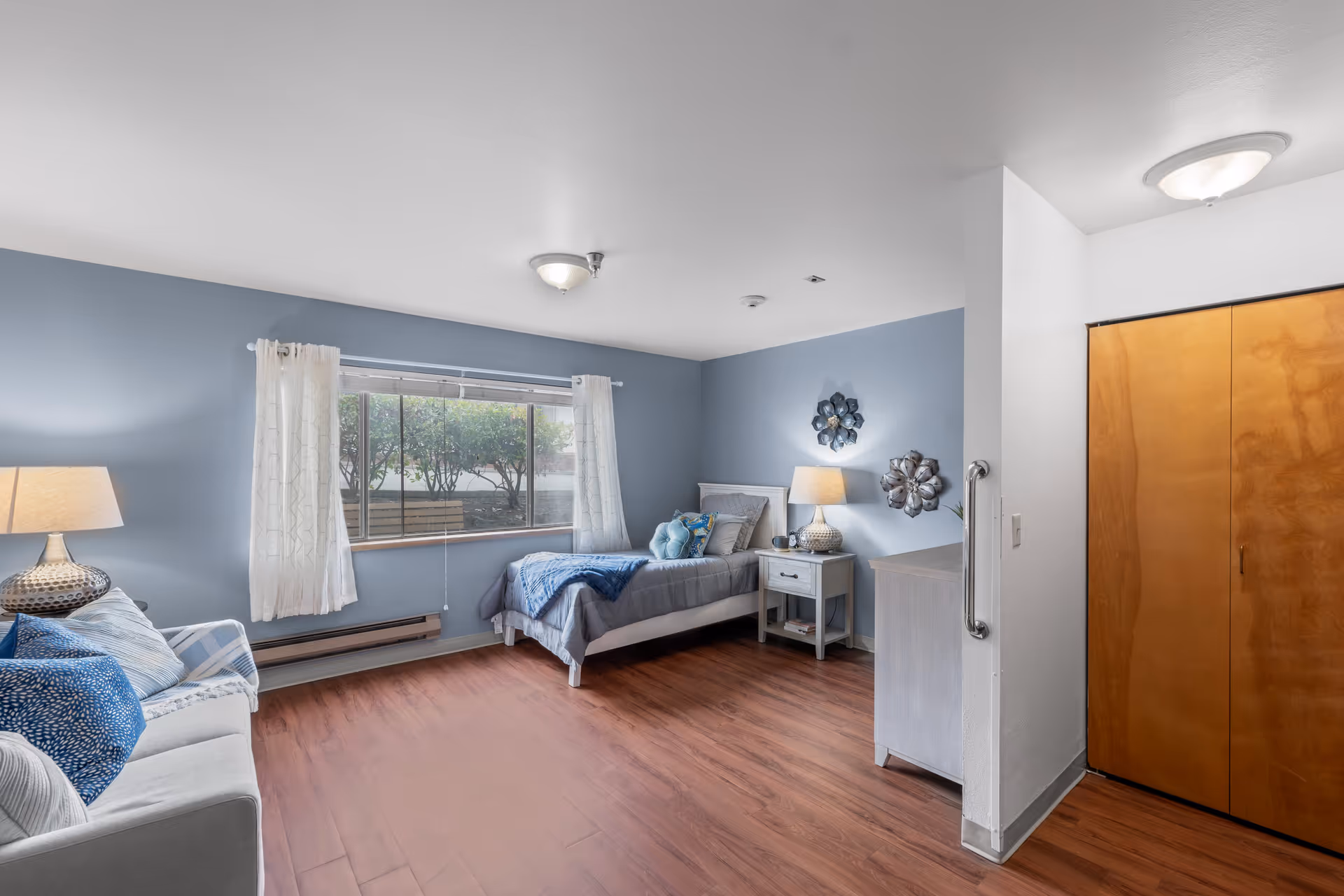 A senior living facility bedroom with light blue walls, a single bed with gray bedding and blue accents, a nightstand with a lamp, a dresser, a sofa with blue and white pillows, a large window with white curtains, and wooden flooring. There is a wooden closet door and a grab bar mounted on the wall near the entrance.