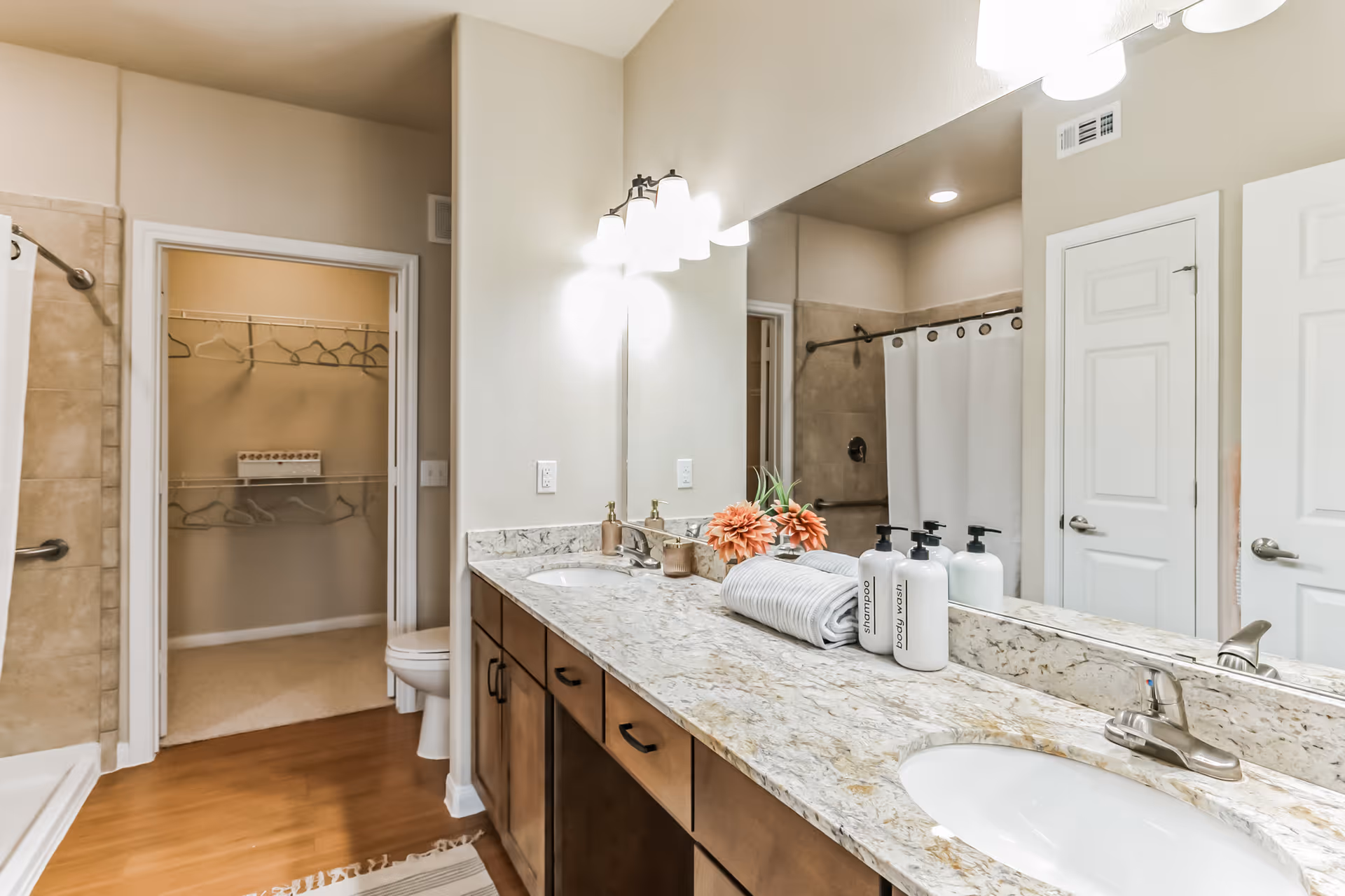 Bright bathroom with a large granite countertop featuring two sinks, a mirror with three light fixtures above, and decorative items including a rolled towel and bottles labeled shampoo and body wash. There is a shower with a white curtain and a walk-in closet with hangers visible through an open door.