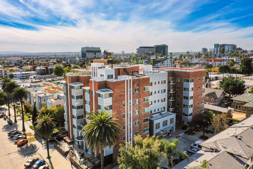 Aerial view of a multi-story brick-and-white senior living building surrounded by palm trees and neighborhood streets.