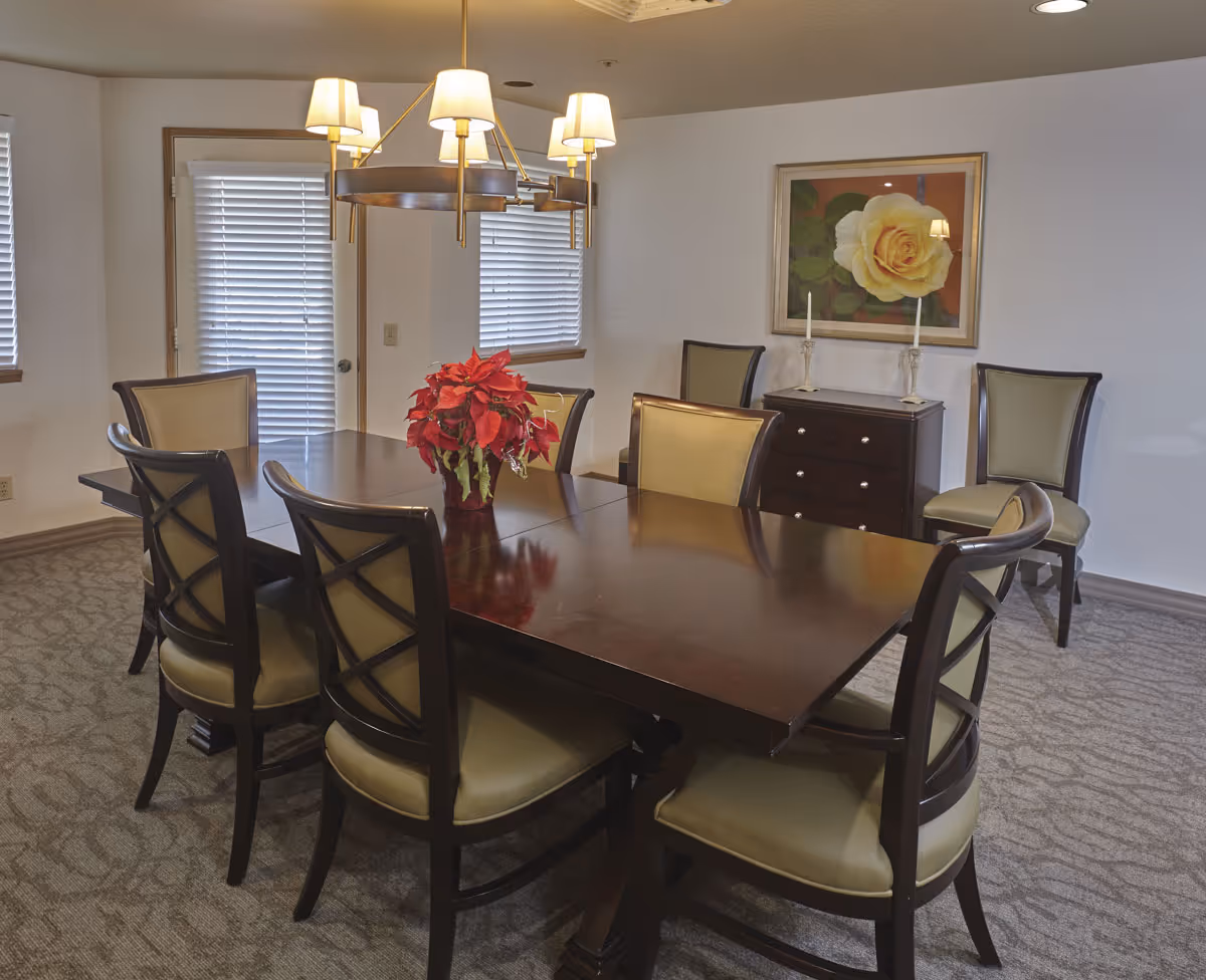 A dining room with a large dark wooden table surrounded by eight chairs with beige cushions. A red poinsettia plant is placed in the center of the table. The room has beige carpet, white walls, two windows with blinds, a door with blinds, a chandelier with multiple lampshades hanging above the table, and a framed picture of a yellow rose on the wall above a small dark wooden cabinet with two candlesticks.