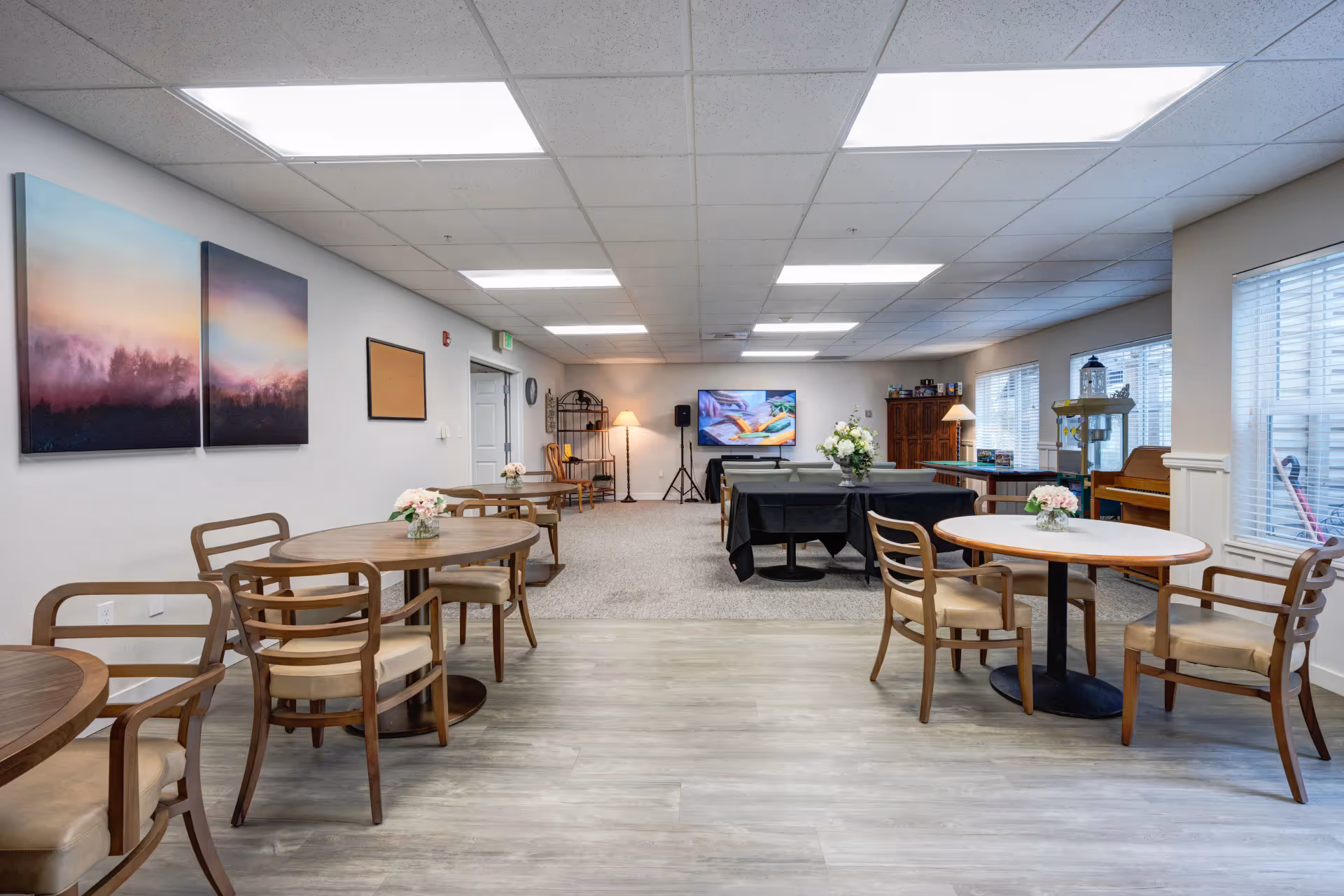 A spacious common area in a senior living facility with several round wooden tables and chairs arranged for seating. The room has light-colored walls with two landscape paintings, large windows with blinds, and a flat-screen TV mounted on the far wall. There are decorative flowers on the tables, a popcorn machine, a piano, and a lamp in the background, creating a welcoming and comfortable atmosphere.