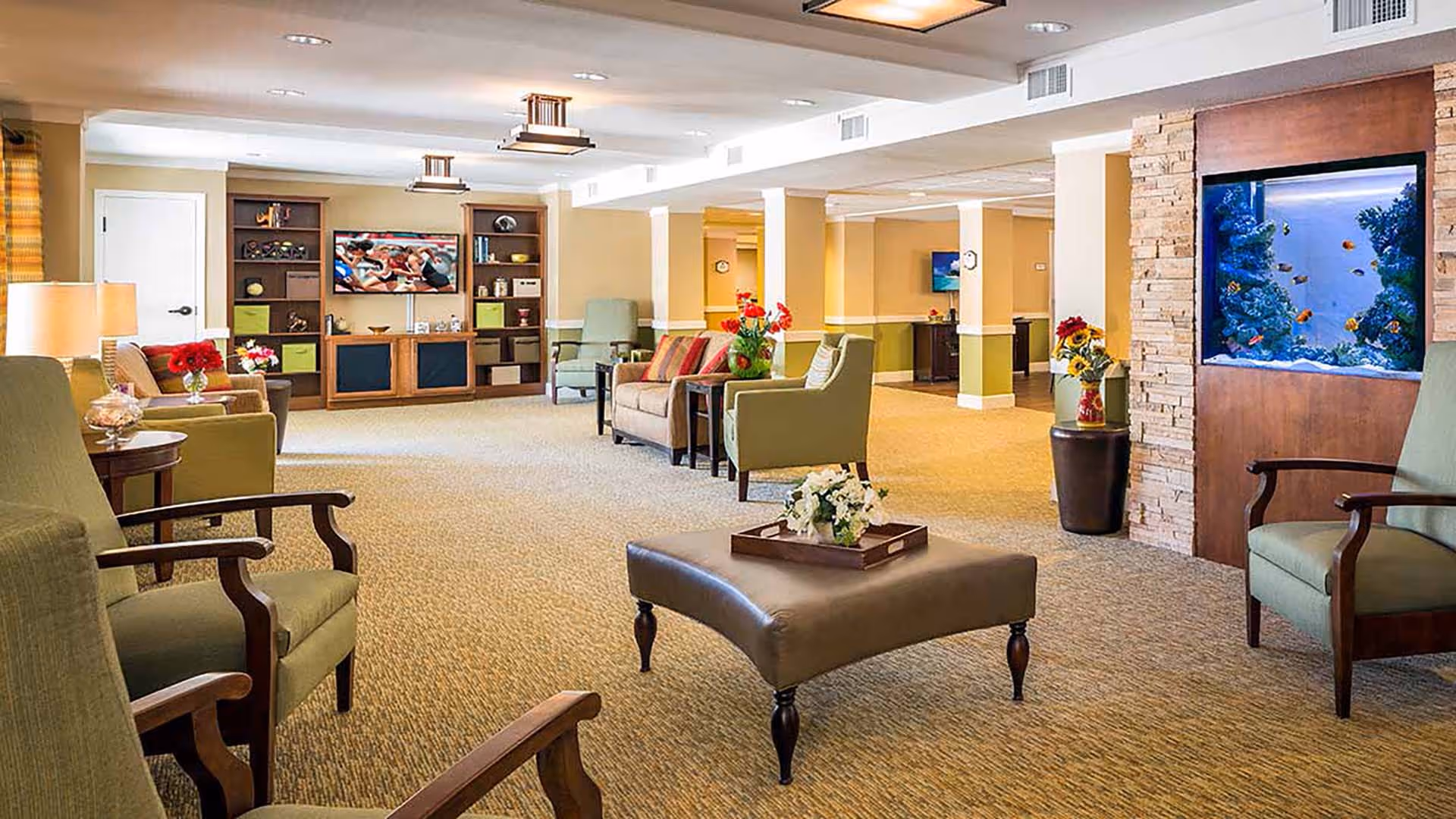 A spacious and well-lit living room area in a senior living facility featuring comfortable armchairs, a central ottoman with a flower arrangement, a large built-in aquarium, and a wall-mounted television with shelving units. The room has beige walls, carpeted flooring, and decorative flowers placed throughout.