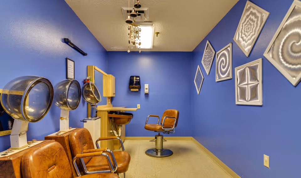 Interior of a salon room with blue walls featuring three vintage hair drying chairs with attached hair dryers on the left side, a single salon chair in front of a mirror and sink on the back wall, and six framed abstract art pieces arranged diagonally on the right wall.