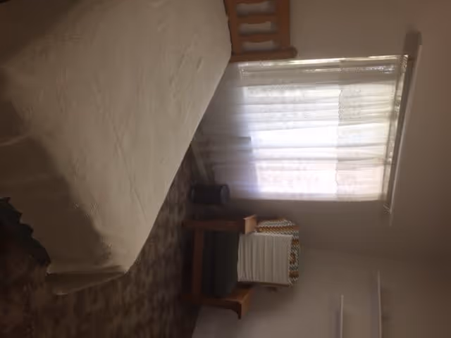 A bedroom with a bed covered in a white quilt, a wooden chair with a cushion and a folded blanket on it, a small trash bin, and a window with sheer white curtains letting in natural light.