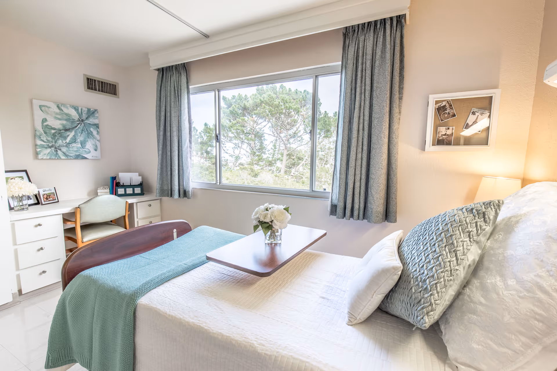 A bright and cozy bedroom in a senior living facility with a neatly made bed featuring white and teal bedding, a small wooden overbed table with a vase of white flowers, a large window with teal curtains showing trees outside, a desk with a chair, framed pictures, and soft lighting from a lamp on the wall.