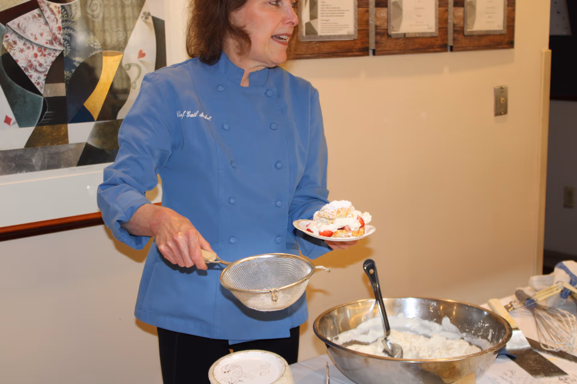 A woman wearing a blue chef's jacket is holding a plate with a dessert topped with whipped cream and strawberries in one hand and a metal sieve in the other. In front of her is a large metal bowl filled with whipped cream and a spoon. The background shows a wall with framed papers and a piece of abstract artwork.