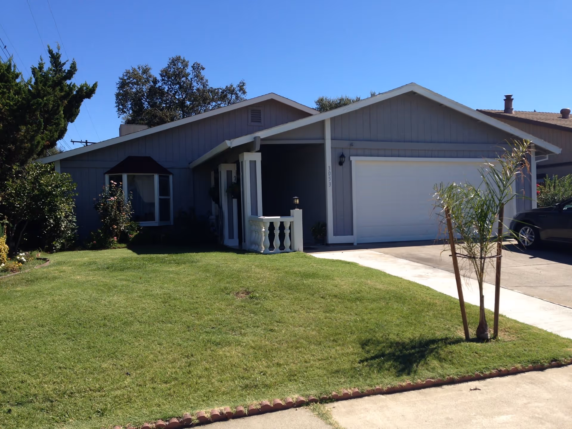 Single-story residential house with a gray exterior, white trim, a two-car garage, and a well-maintained front lawn with a small tree and bushes under a clear blue sky.