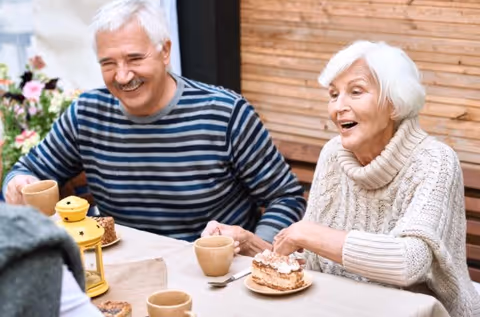 An elderly man and woman sitting at a table outdoors, enjoying coffee and cake together. The man is wearing a striped sweater and smiling, while the woman in a cream-colored sweater looks pleasantly surprised or engaged in conversation. There is a small yellow lantern on the table and flowers in the background.