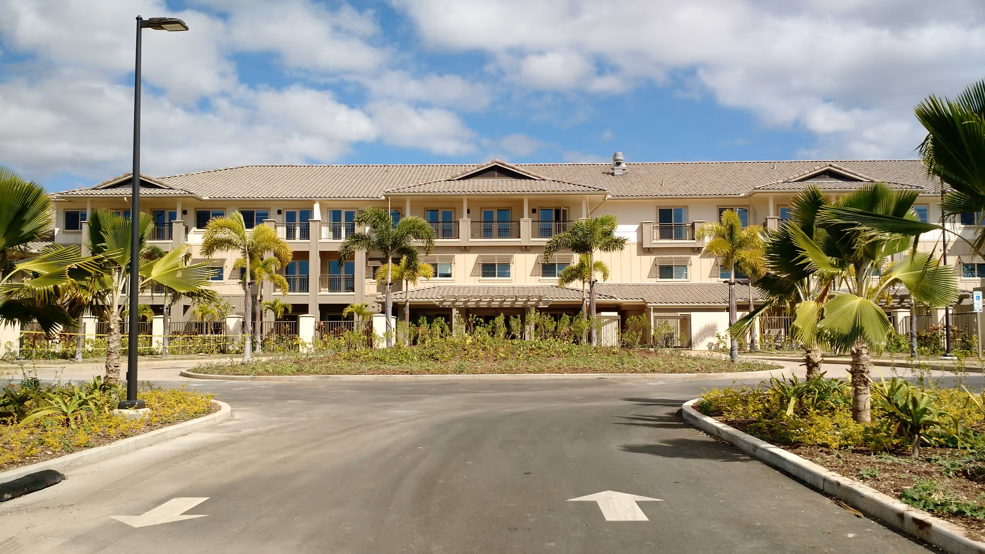 Front exterior view of a three-story senior living facility named 'Ilima at Leihano' with a tiled roof, multiple windows, palm trees, and a driveway with directional arrows under a partly cloudy sky.