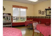 Interior view of a dining room in a senior living facility with tables covered in red tablecloths, chairs with striped upholstery, a window with blinds and a valance, framed pictures on the wall, and a small cabinet with decorative items.