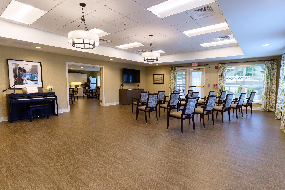 A spacious senior living common area with rows of chairs arranged facing a wall-mounted television. The room features two large ceiling light fixtures, a piano with a music stand on the left side, and large windows with floral curtains allowing natural light to enter. Through an open doorway, a kitchen or dining area with tables and chairs is visible.